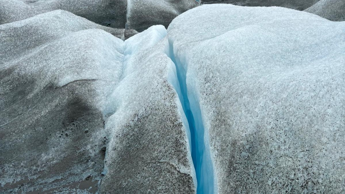 A glacier surface split by a vivid blue crevasse, with pale ice and dust-covered ridges stretching toward the horizon.