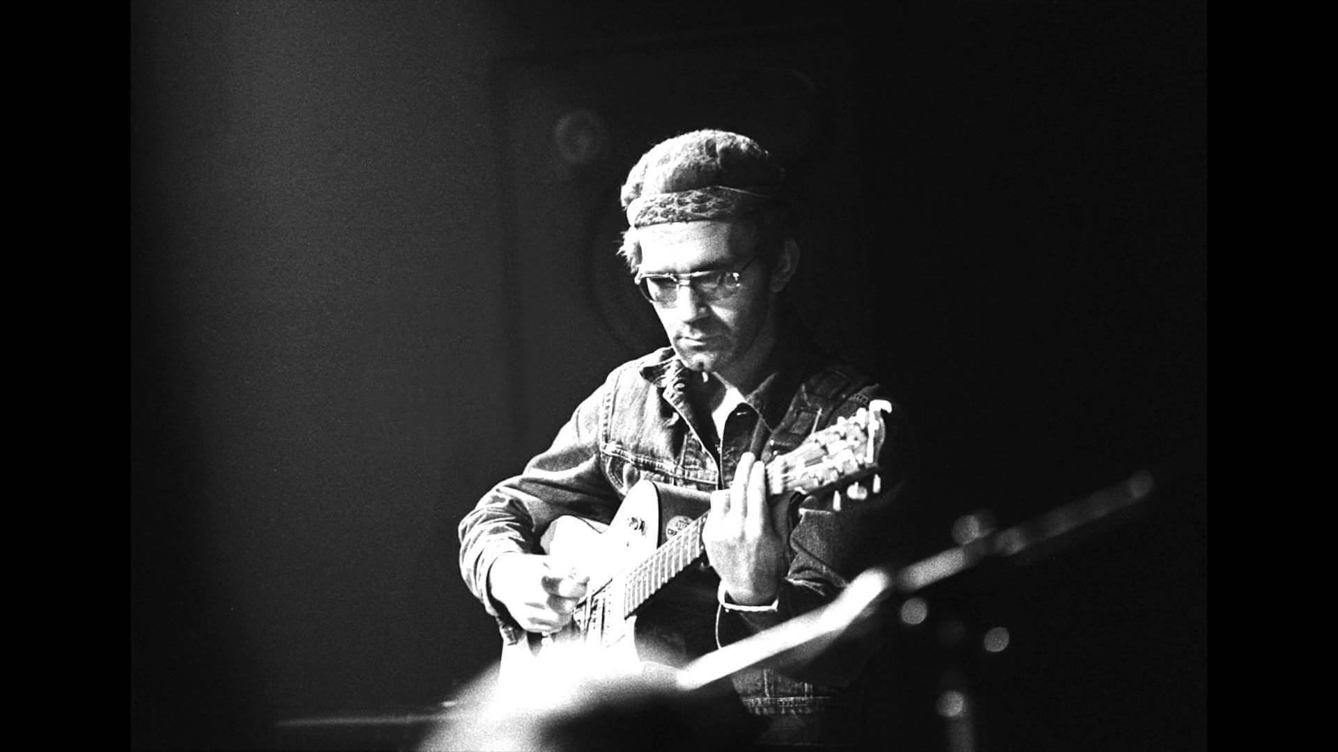 Black-and-white photograph of J.J. Cale performing live on stage, wearing a denim shirt and headband with an electric guitar.